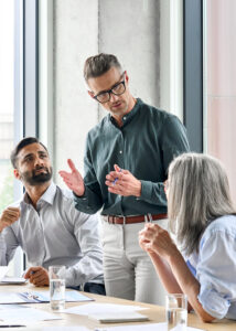 Mature businessman leader mentor talking to diverse colleagues team listening to caucasian ceo. Multicultural professionals project managers group negotiating in boardroom at meeting.
